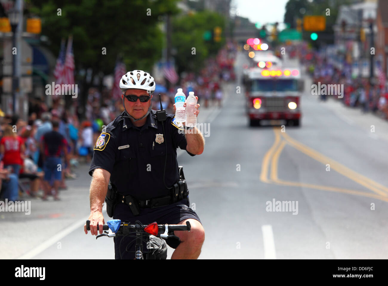Policeman on bicycle holding water bottles to warn against risks of dehydration during 4th July Independence Day parades, Catonsville, Maryland, USA Stock Photo