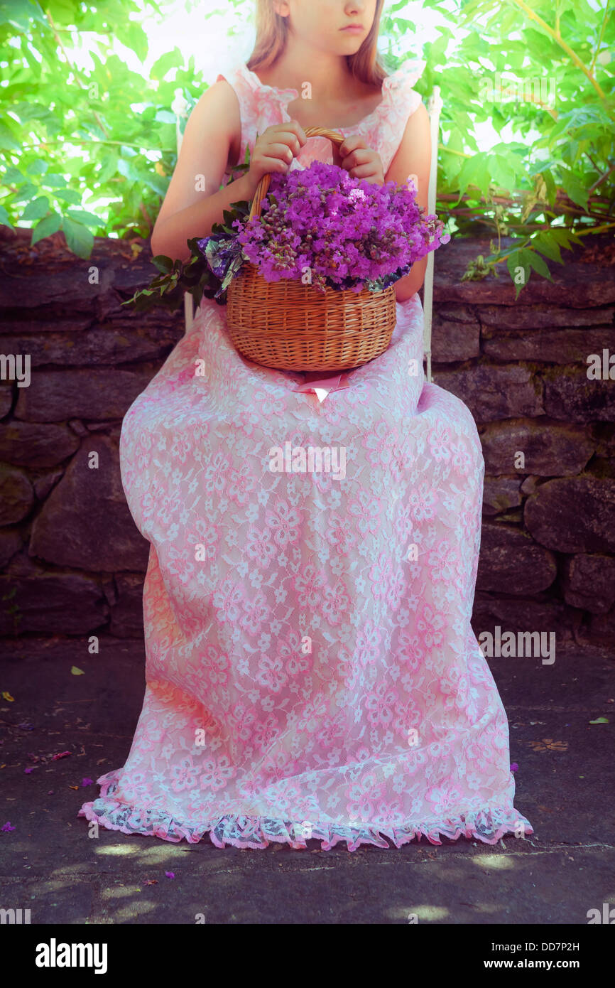 a girl in a pink dress sitting outside on a chair with a basket full of wild flowers Stock Photo
