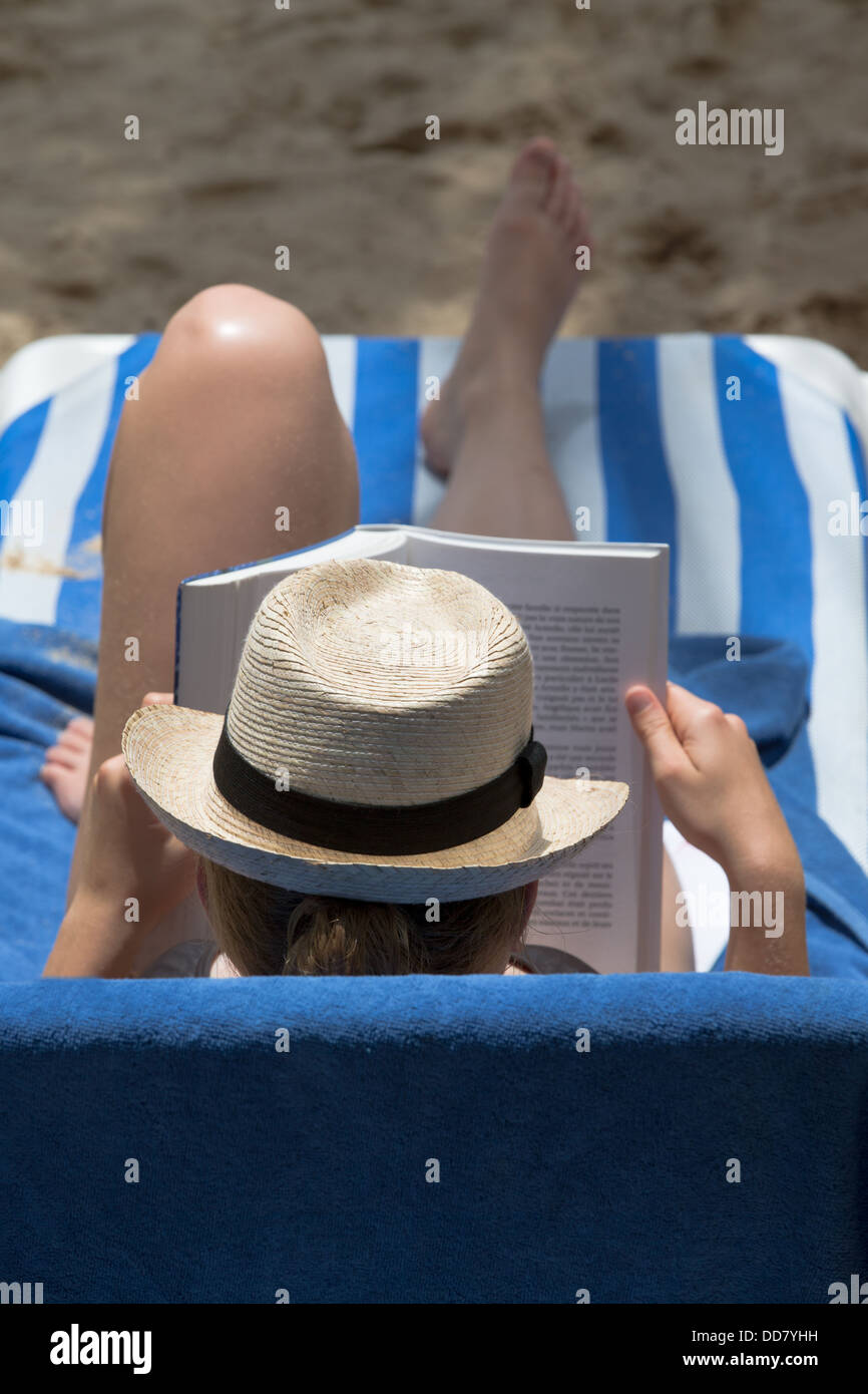 Lady in hat reading her book on the beach recliner Stock Photo