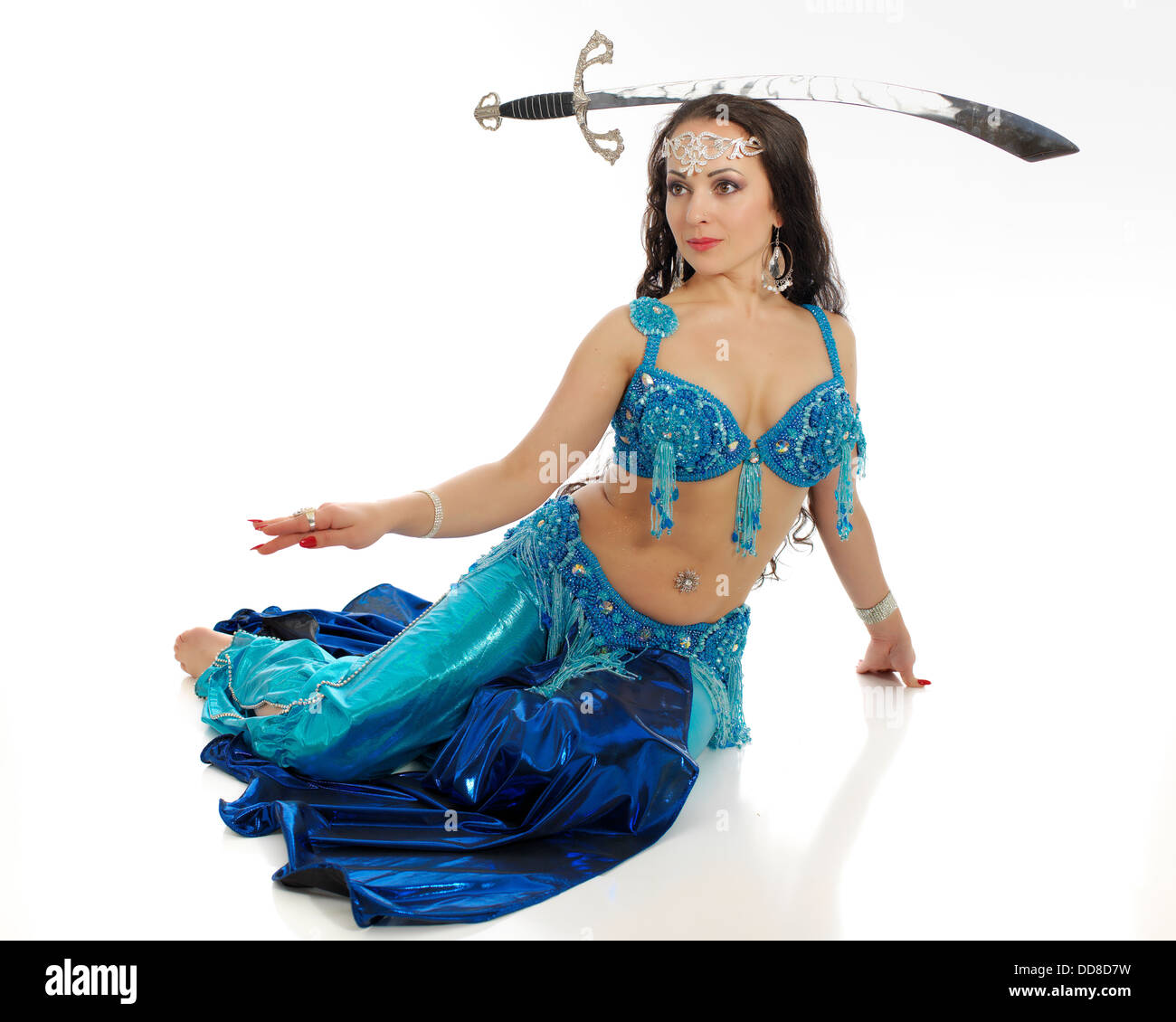Beautiful professional bellydancer  in colorful costume posing in front of the white backdrop with a sward. Stock Photo