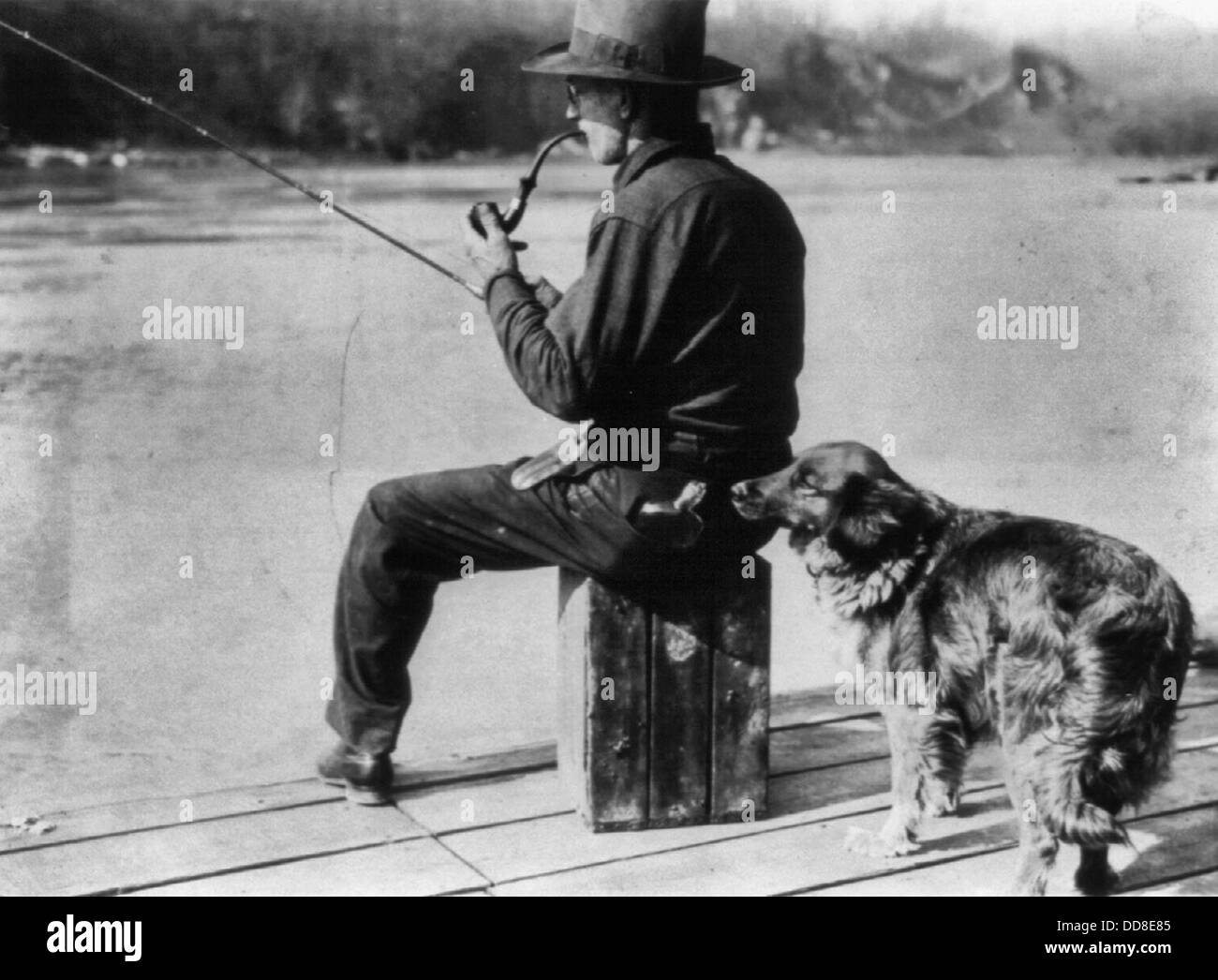 Hooch Hound, a dog trained to detect liquor, sniffs at flask in back pocket of man, seated, fishing on pier on the Potomac River, 1922, Prohibition America Stock Photo