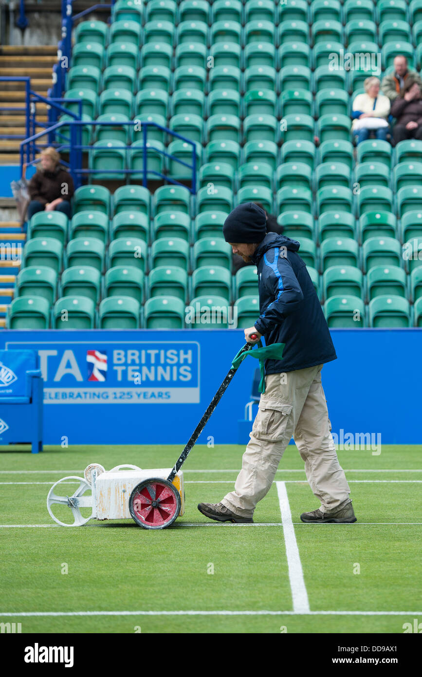 Groundsman paints the white lines on a grass tennis court with a machine as spectators watch on from the stands Stock Photo