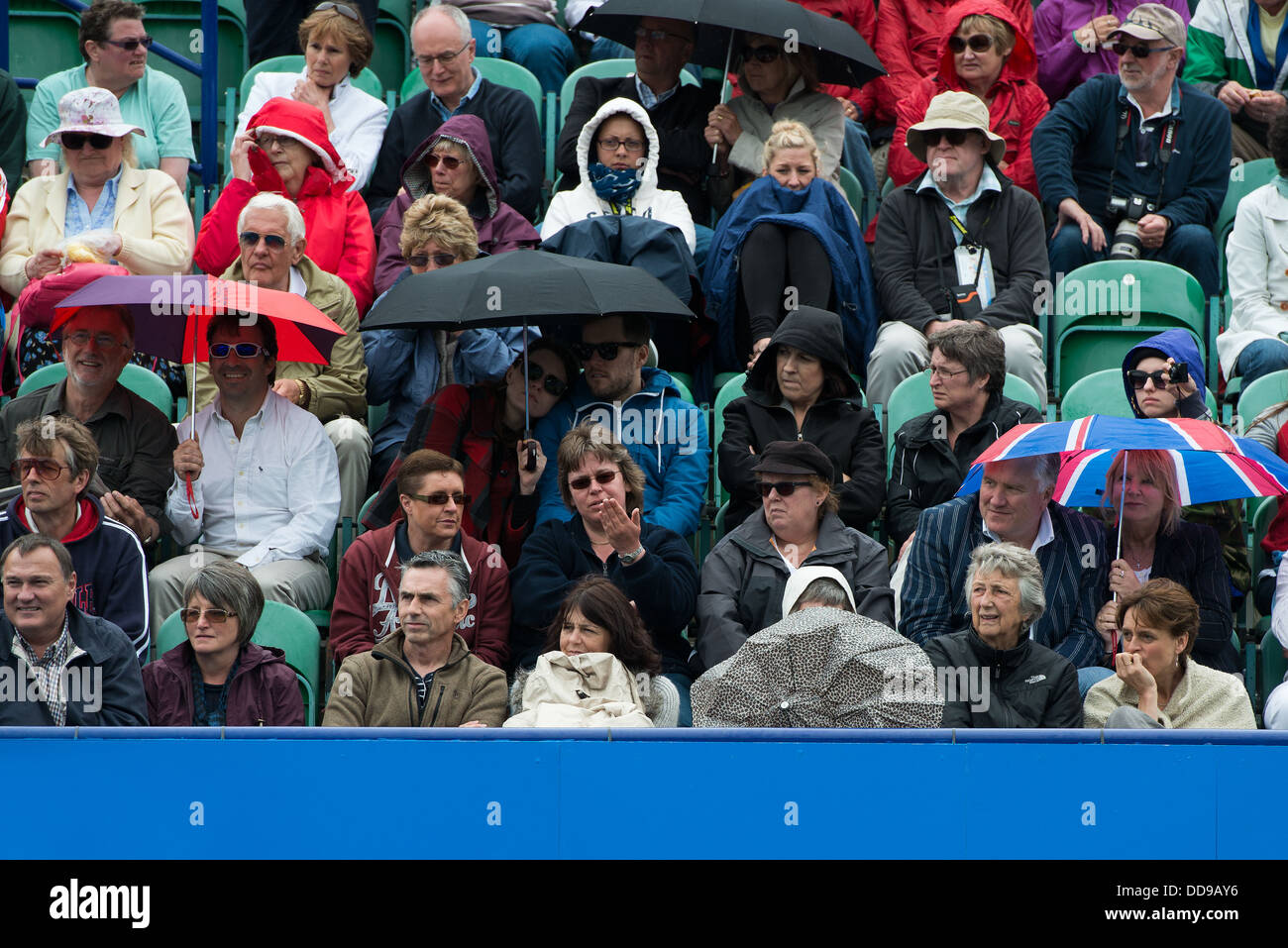 A crowd of spectators at a tennis match shelter in rain jackets and under umbrellas during a break in play Stock Photo