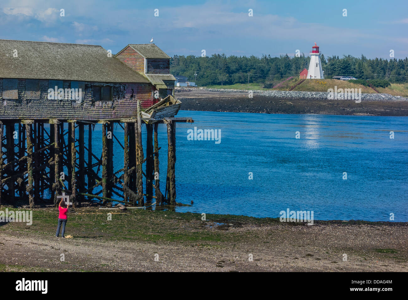 A female artist uses a cropping tool to find the best view of a fish processing building on stilts & lighthouse for a painting. Stock Photo
