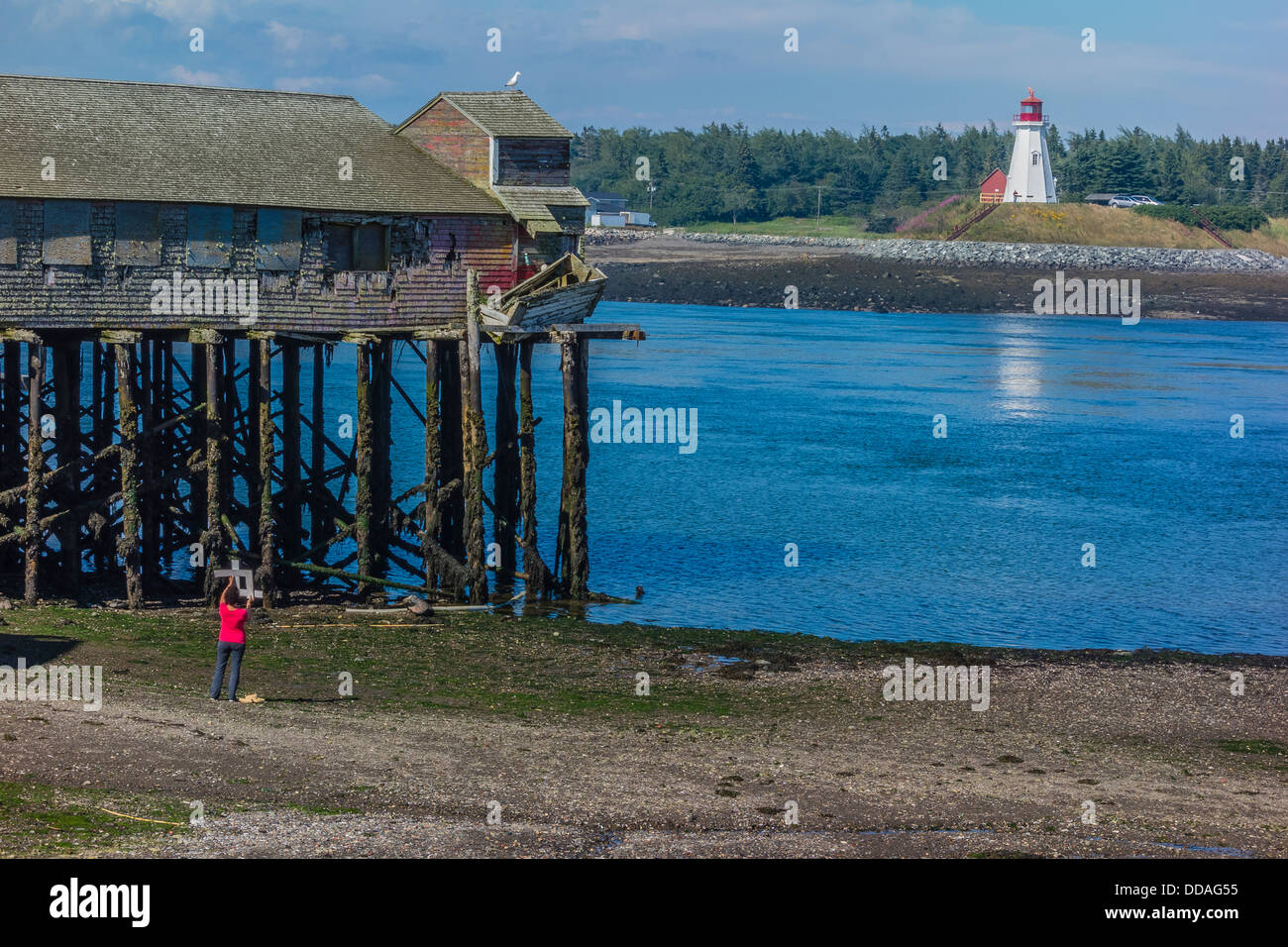 A female artist uses a cropping tool to find the best view of a fish processing building on stilts & lighthouse for a painting. Stock Photo