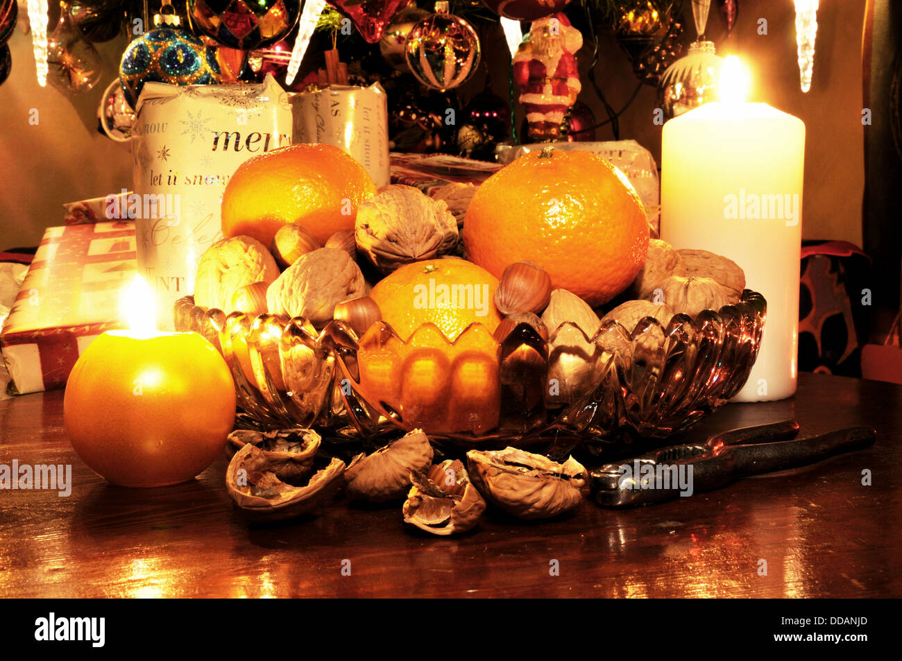 Glass bowl filled with walnuts, hazelnuts and mandarin oranges with Christmas presents under tree to the rear. Stock Photo