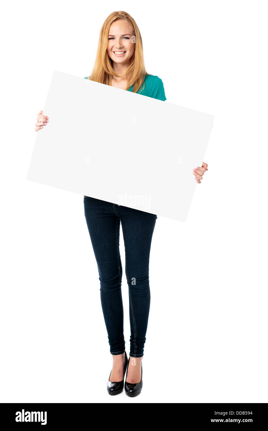 Attractive smiling slender young woman holding a blank white rectangular sign at an angle in front of her body, full length on white Stock Photo