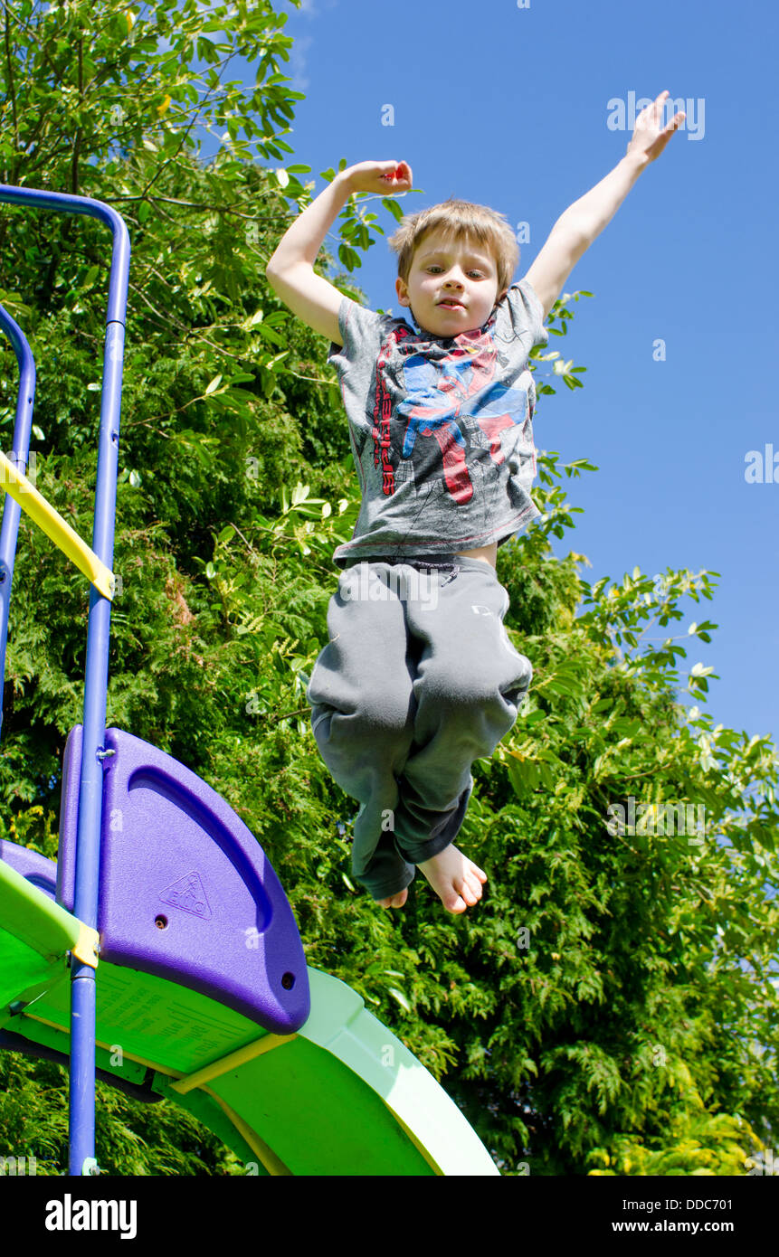 eight year old boy jumping from a climbing frame slide in garden onto a lawn. Stock Photo