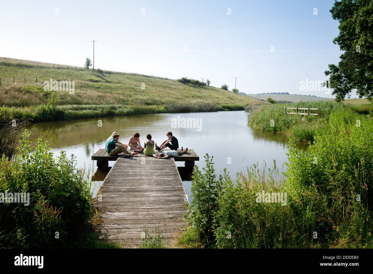 Young people sitting on a jetty by a lake in english countryside, Sheepdrove Organic Farm, Lambourn, Berkshire, UK Stock Photo