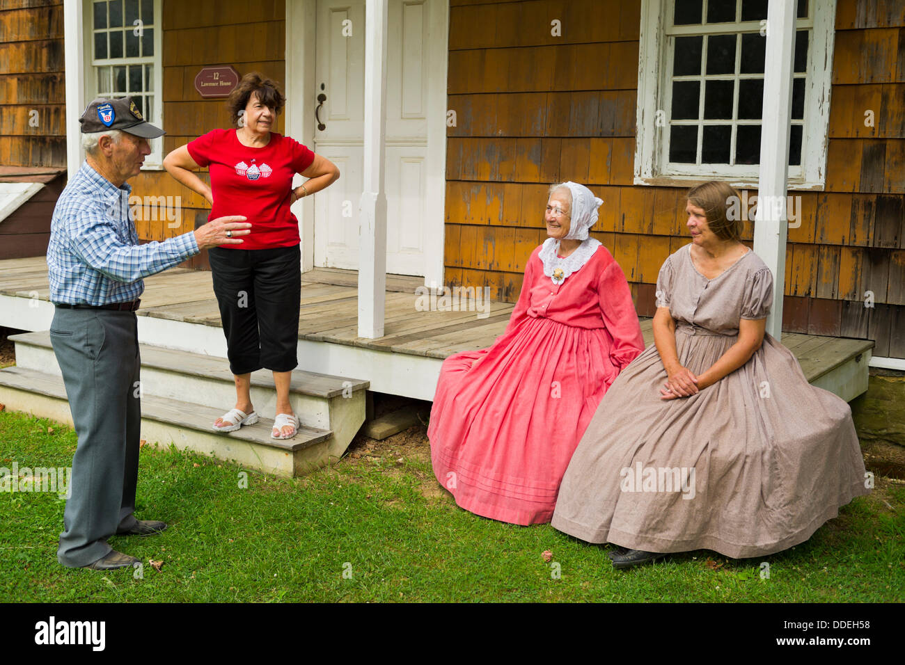 Old Bethpage, New York, U.S. 31st August 2013. PATRICIA JOSEPH, of College Point, in red dress, and JANE HEILIG, of Bethpage, in taupe dress, are talking with a man and woman visiting during the Olde Time Music Weekend at Old Bethpage Village Restoration, where 1800's popular music and dances of the American Civil War period is performed. Joseph & Heilig, wearing American Civil War era style clothing and sitting on the porch of 12 Lawrence House, are members of the Old Bethpage Village Dancers. Stock Photo