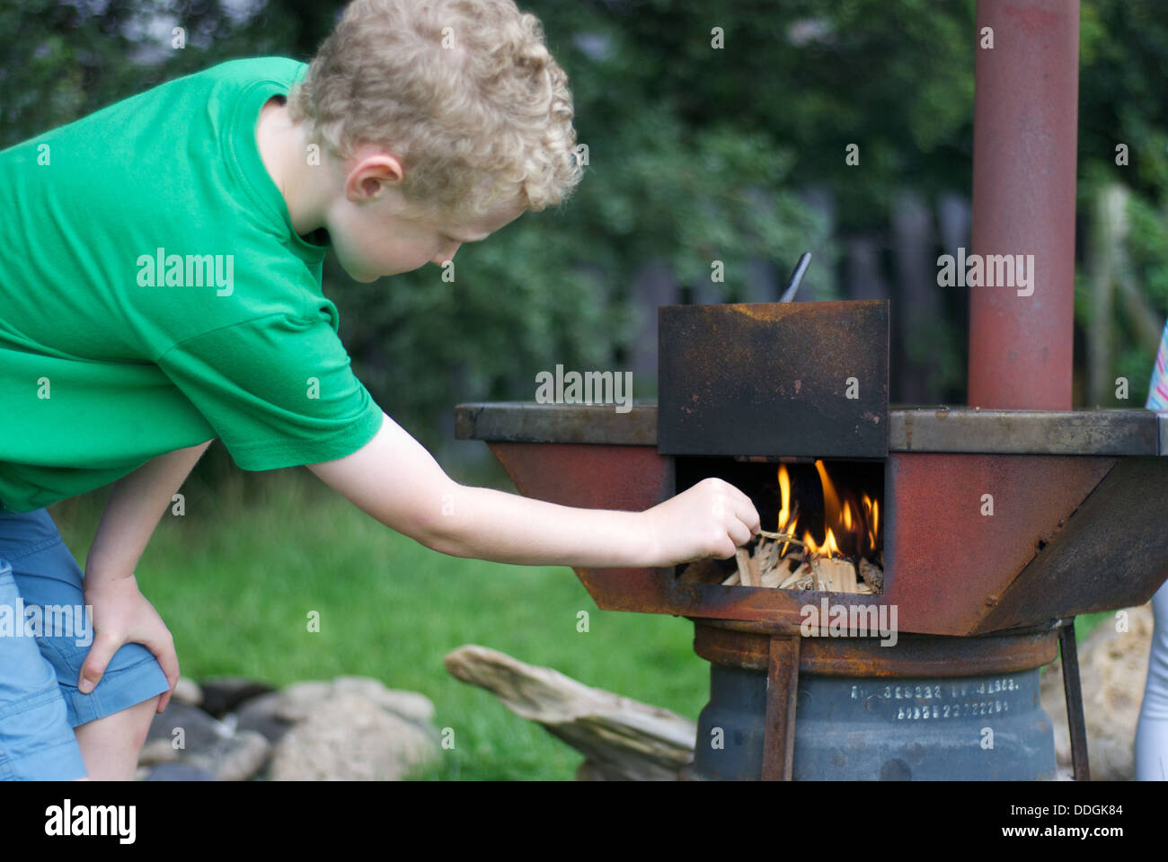 A young blond boy lighting a fire in an outdoor wood burning stove on a campsite Stock Photo