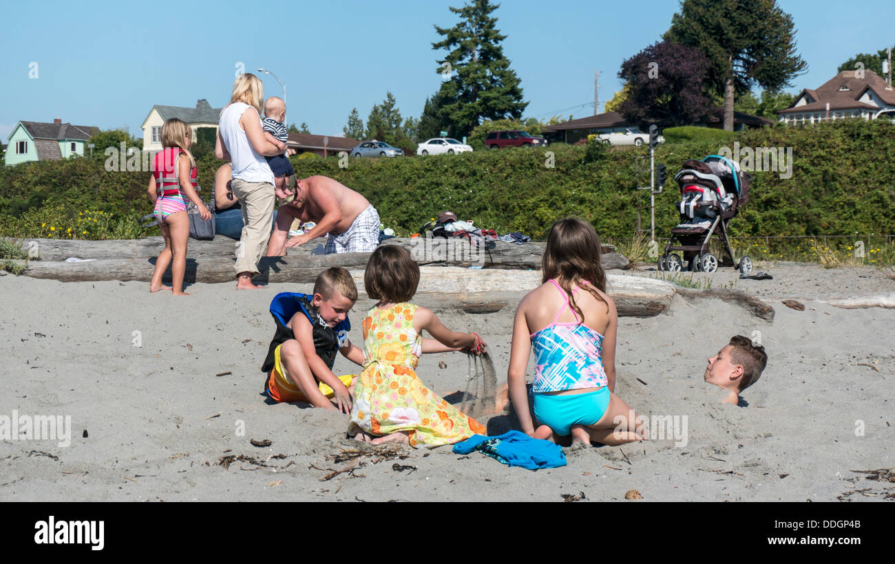 2 little girls bury a bigger boy in the sand while small brother looks on & parents watch baby Edmonds waterfront beach Stock Photo