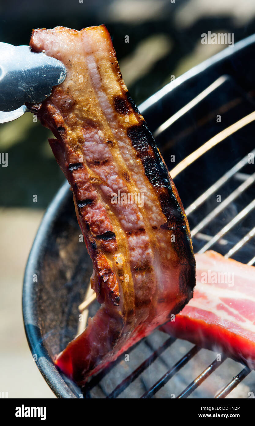 Two thick pieces of bacon on grill.One is being picked up with tongs Stock Photo