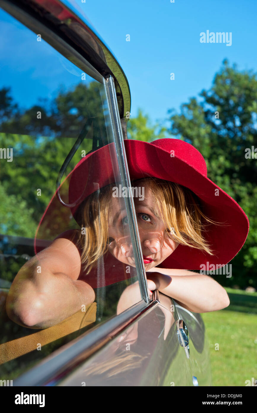 Young woman with a large red felt hat leaning out of the window of a French oldtimer on a bright, sunny, summer day Stock Photo