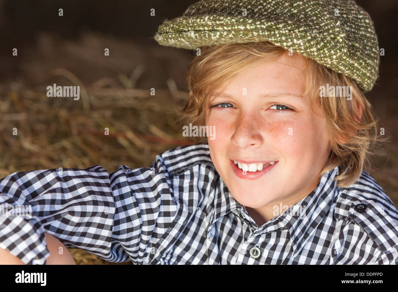 Young happy smiling blond boy child aged twelve or early teenager wearing  plaid shirt & flat cap sitting on hay or straw bales Stock Photo