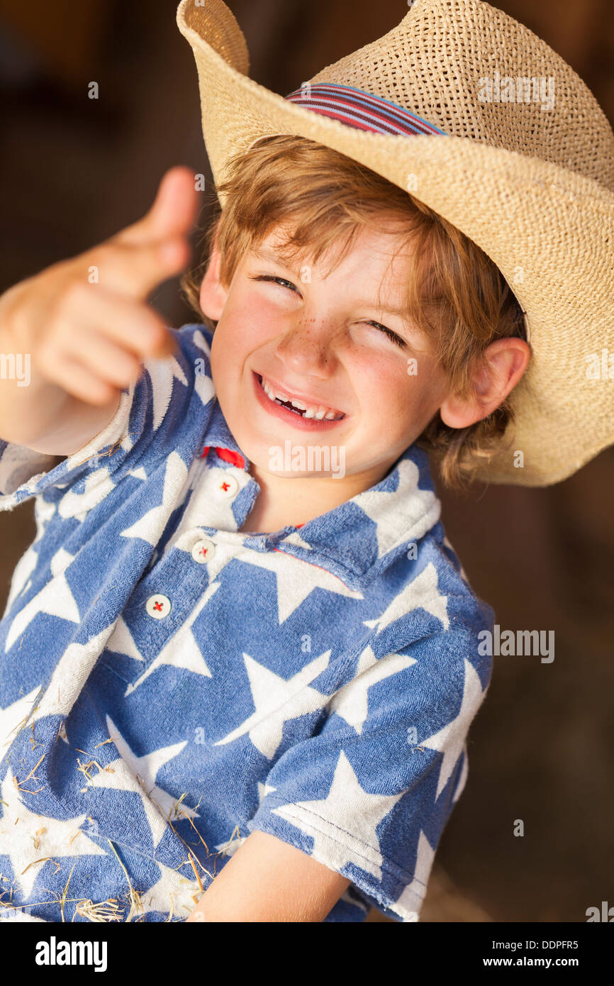 Young happy smiling blond boy child wearing a blue star shirt and cowboy hat sitting on hay or straw bales making a finger gun Stock Photo
