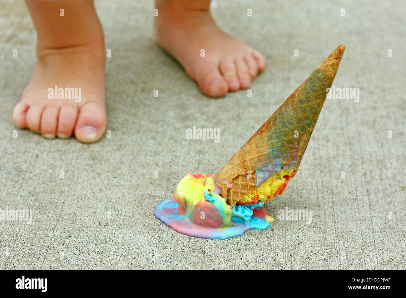 a dropped rainbow colored ice cream cone lays upside down on the sidewalk at the feet of a young child Stock Photo