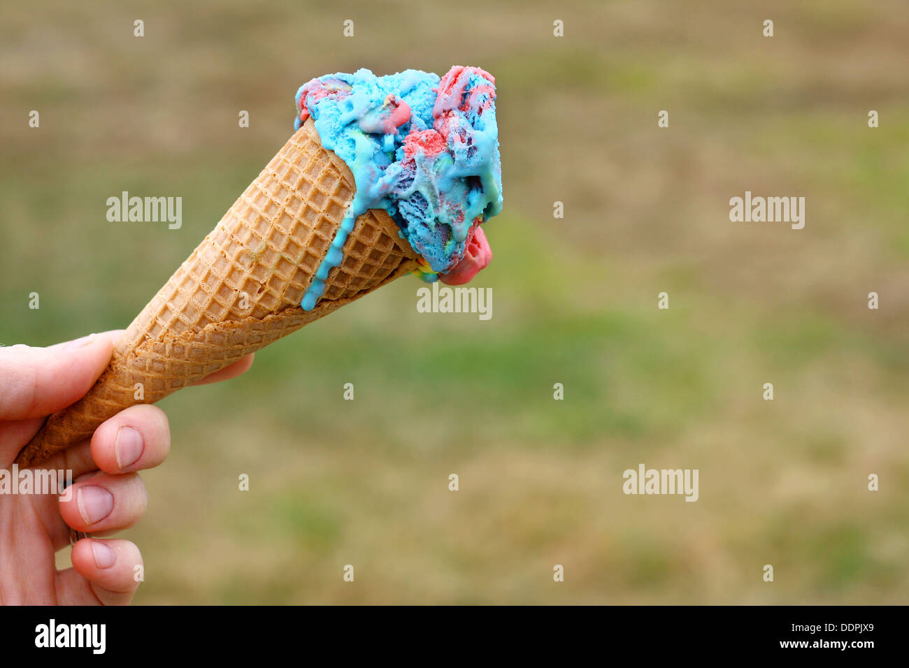 a hand is holding a blue and pink cotton candy flavored ice cream cone in front of a  green grass background on a summer day Stock Photo