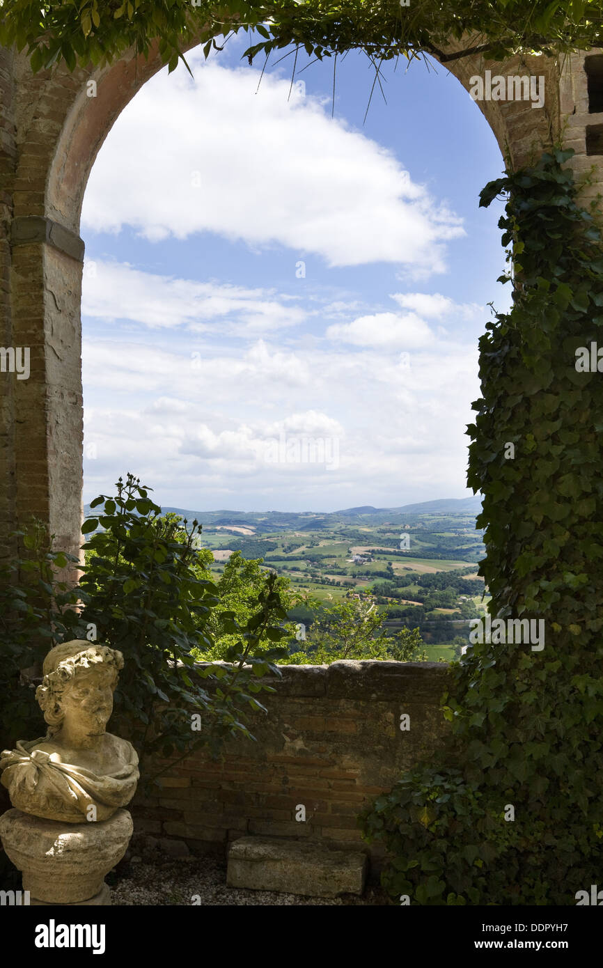 View over Umbrian landscape as seen through arch of garden courtyard, surrounded by trailing and ivy, and stone statue, Renaissa Stock Photo