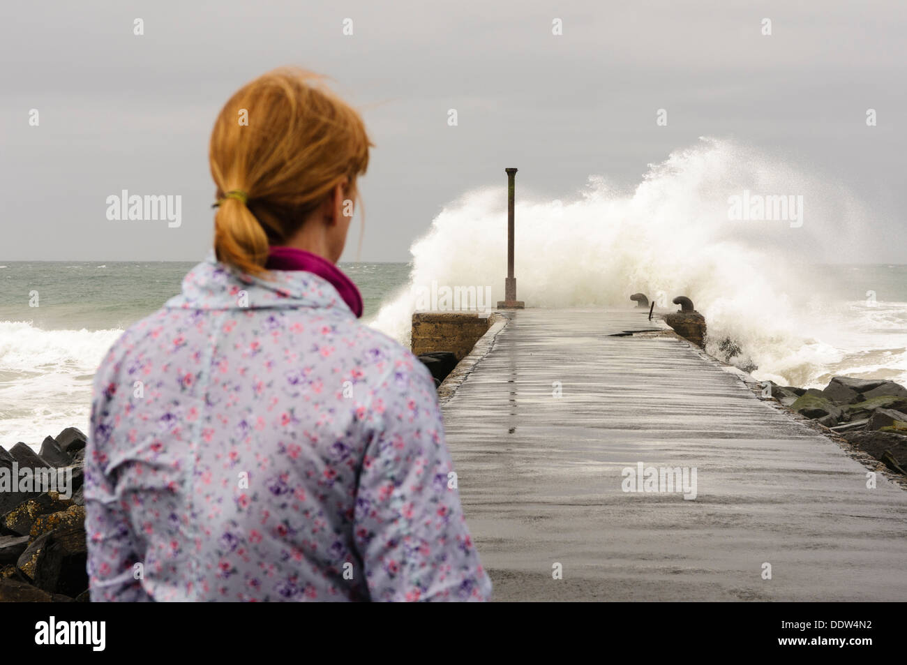 A woman watches as a wave crashes over the end of a pier in windy conditions Stock Photo