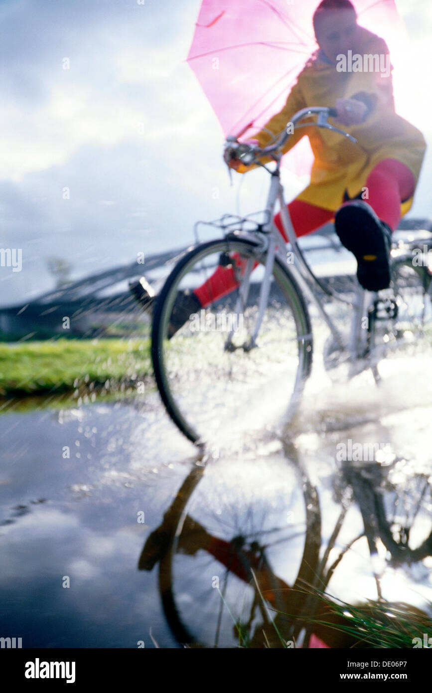 Cyclist with a raincoat and an umbrella riding through a puddle of water Stock Photo