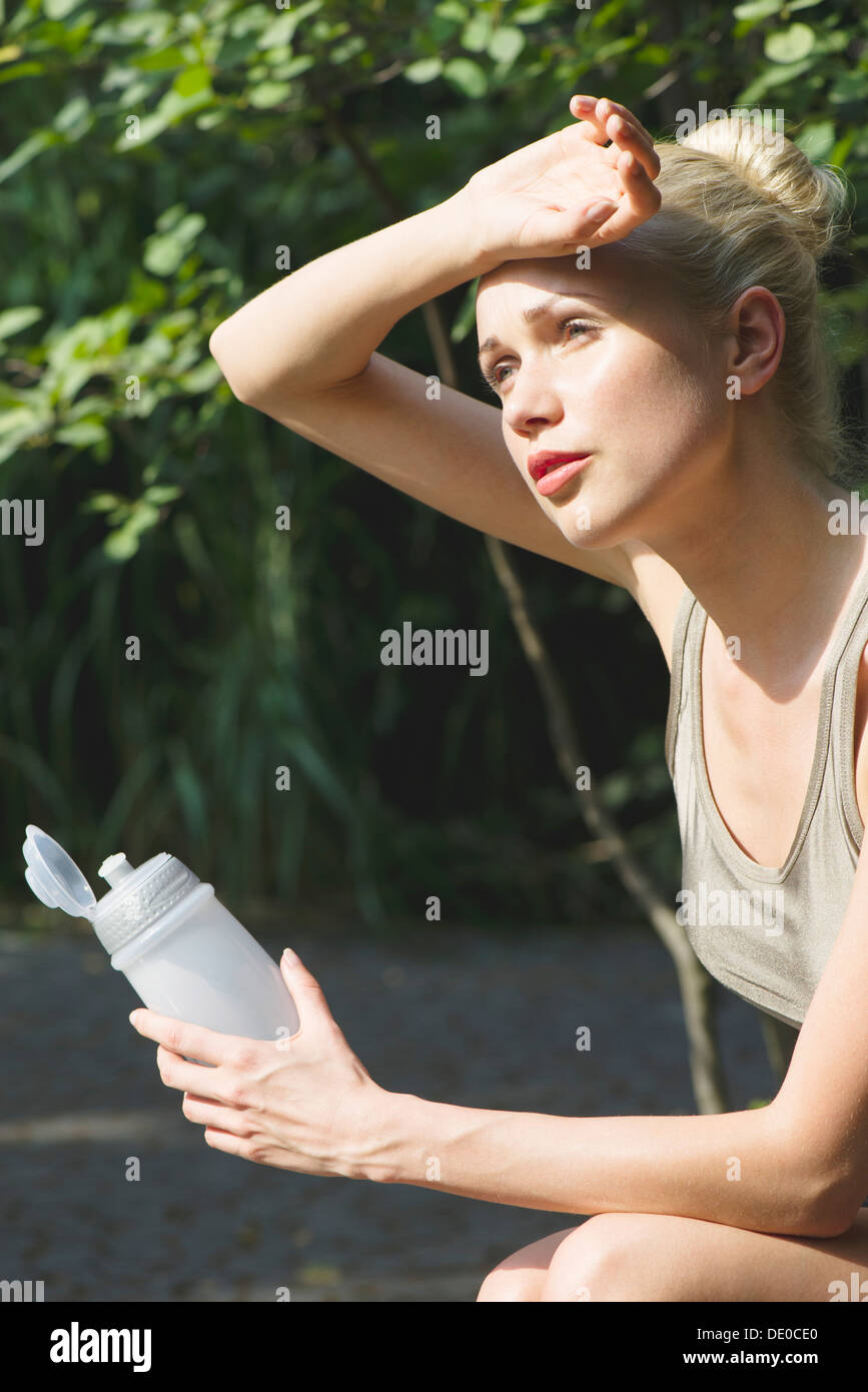 Young woman sitting outdoors with water bottle, wiping forehead with back of hand Stock Photo