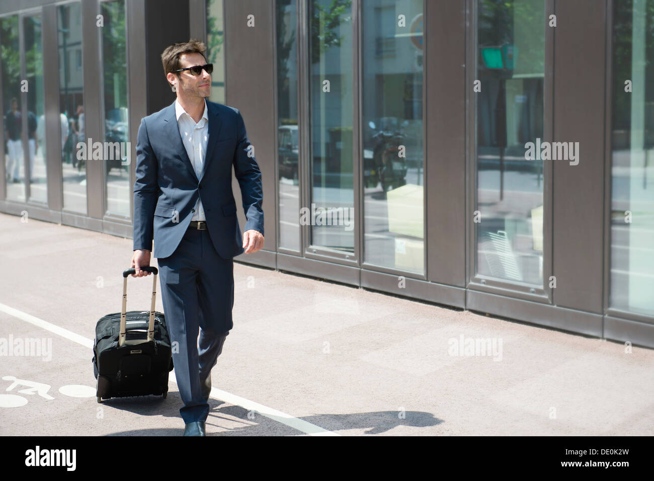 Businessman with sunglasses pulling luggage, walking in city Stock Photo