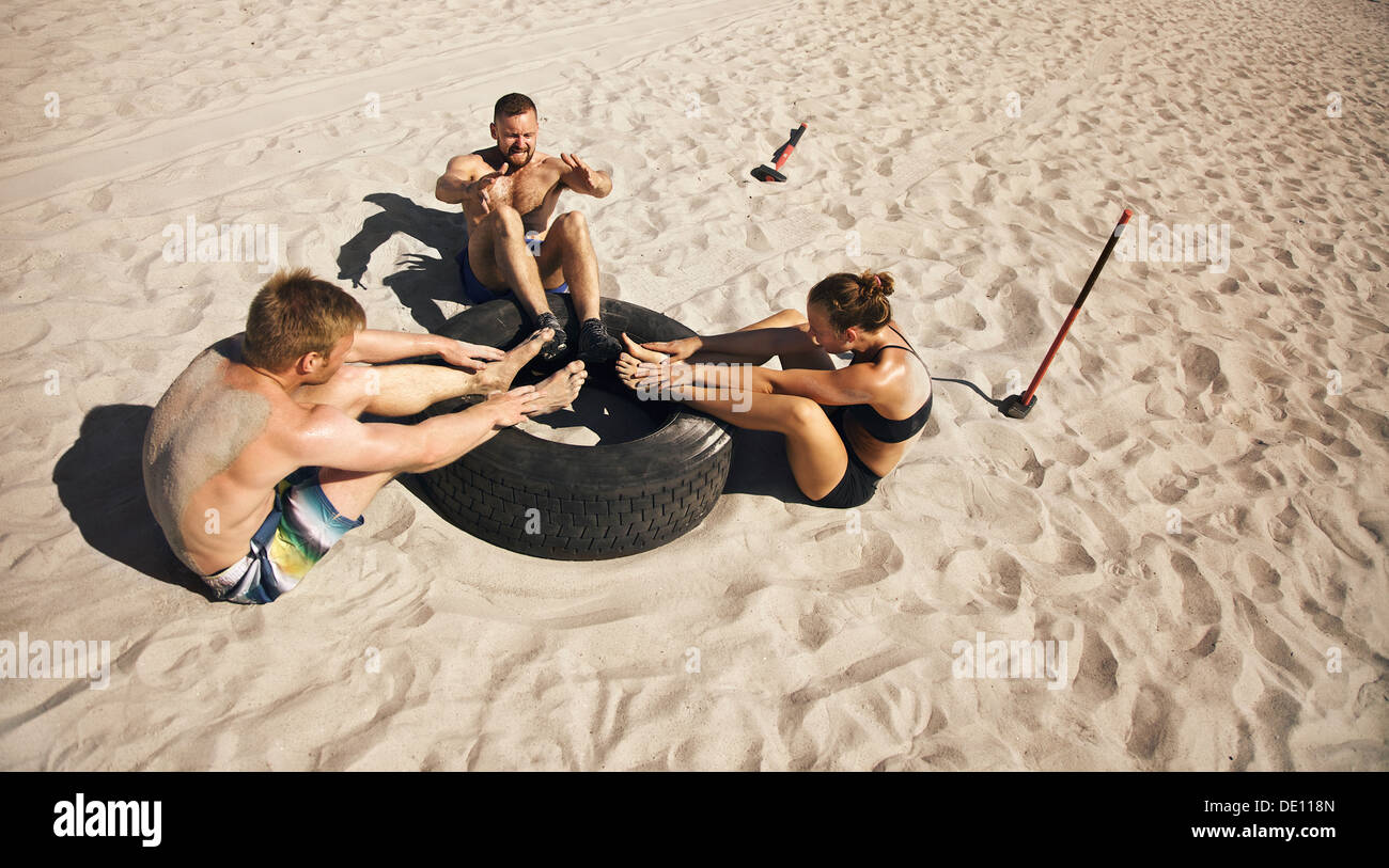 Fitness and healthy lifestyle. Small group of young athletes doing abdominal exercise with a truck tire on beach. Athletes doing Stock Photo