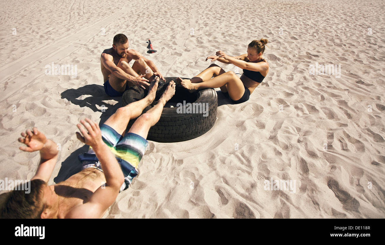 Three young athletes doing abdominal exercise with a truck tire on beach. Athletes doing crossfit workout outdoors Stock Photo
