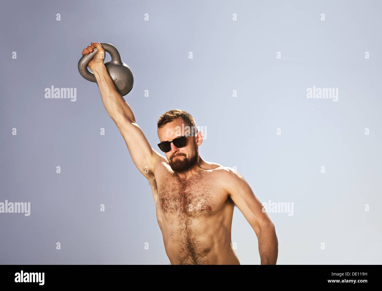 Male athlete swinging a kettle bell over his head with one hand. Young hipster man doing crossfit workout on a hot summer day. Stock Photo