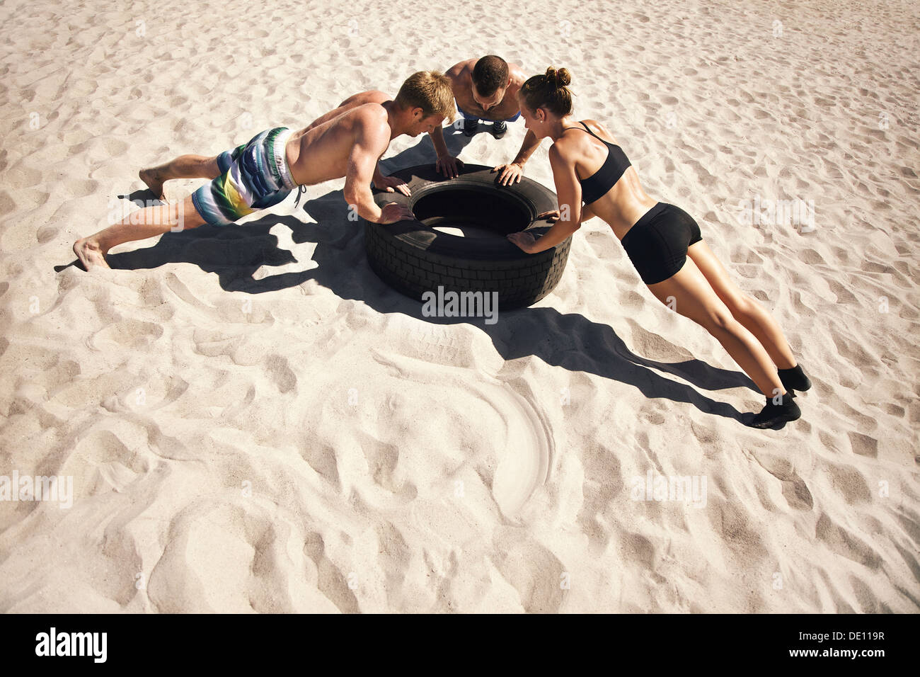 Small group of people doing push-ups on tire. Young athletes working out on beach during a hot summer day. Stock Photo