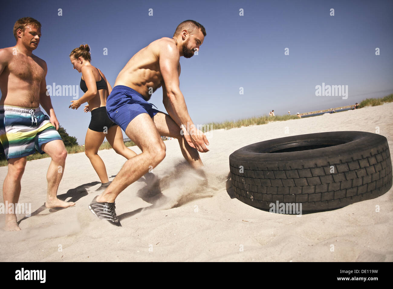 Strong male athlete about to flip a truck tire. Young people doing crossfit exercise on beach on a sunny day. Stock Photo