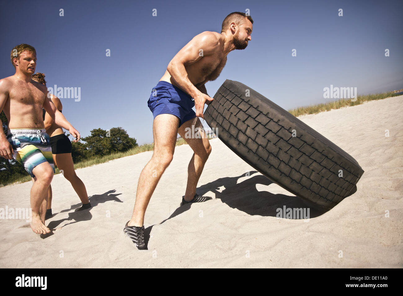 Tough male athlete flipping a truck tire. Young people doing crossfit exercise on beach. Stock Photo