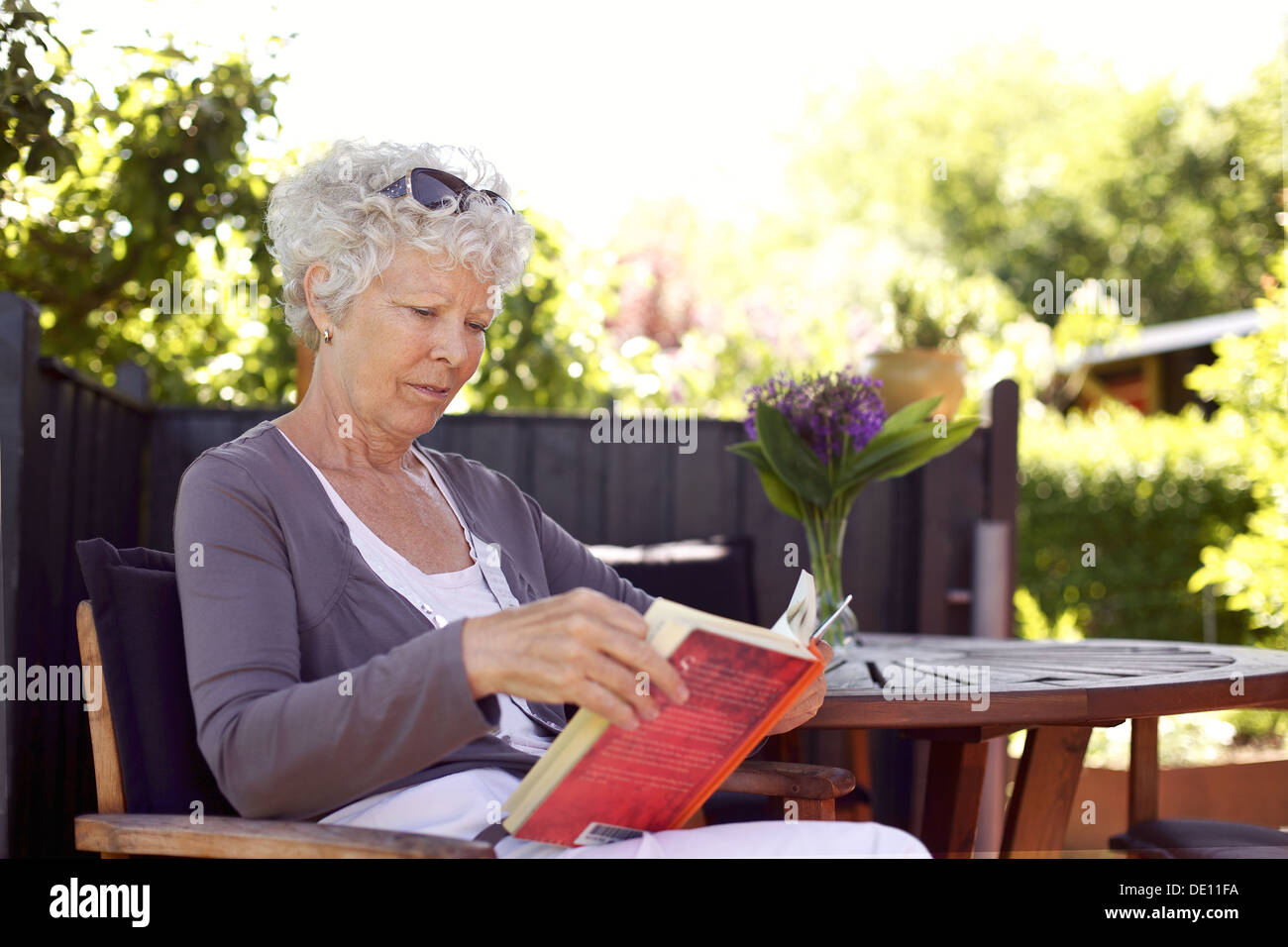 Relaxed senior woman sitting a chair in backyard garden reading a book ...