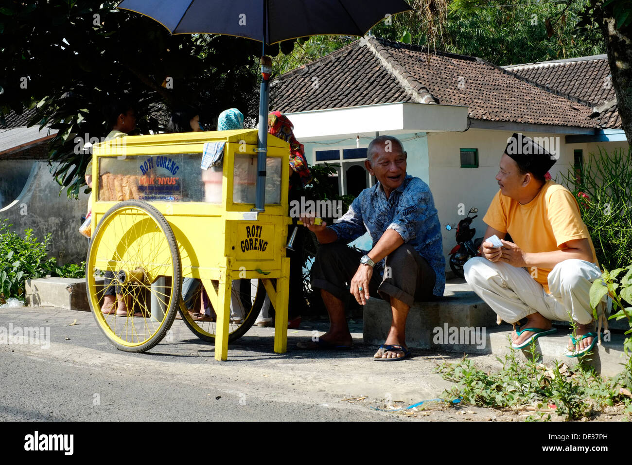 local vendor with his snack cart chats to parents as he waits for children to leave school and buy from him java indonesia Stock Photo