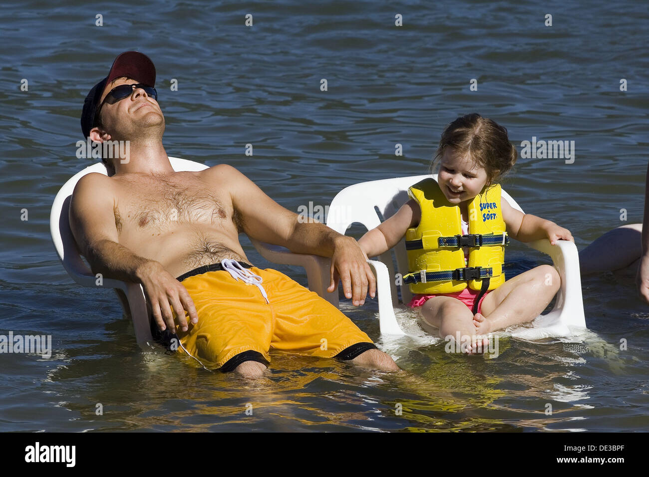 A man, 20-30, and a little girl cool off sitting in chairs in a lake. Stock Photo