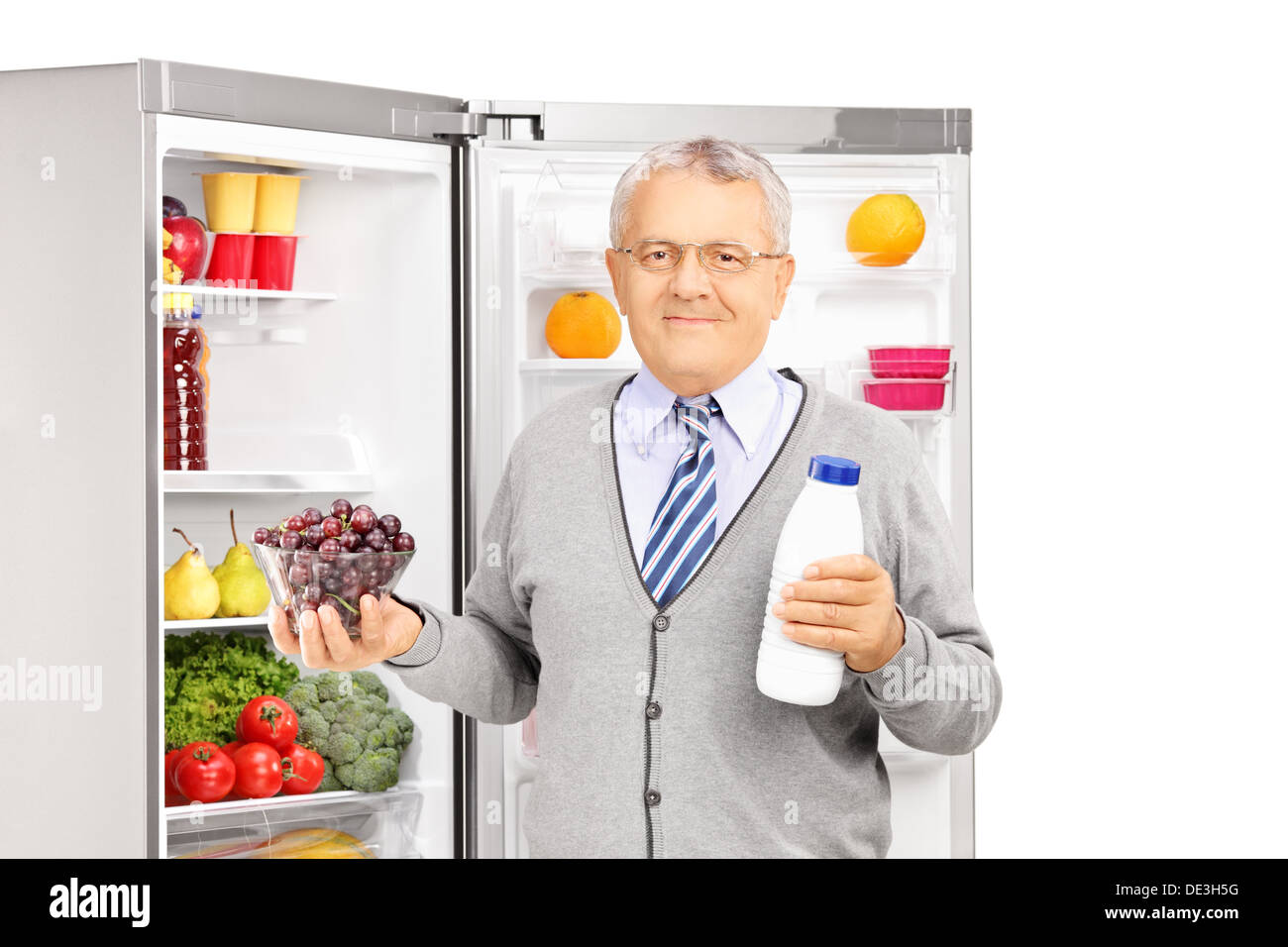 Smiling mature man holding a milk bottle and bowl of grapes next to a refrigerator Stock Photo