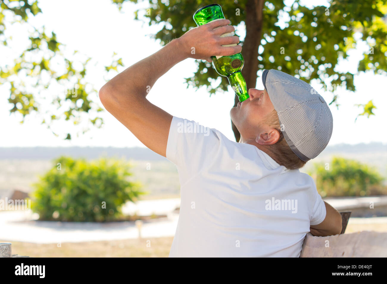 Rear view of an alcoholic man drinking from a bottle of white wine in the park in summer Stock Photo
