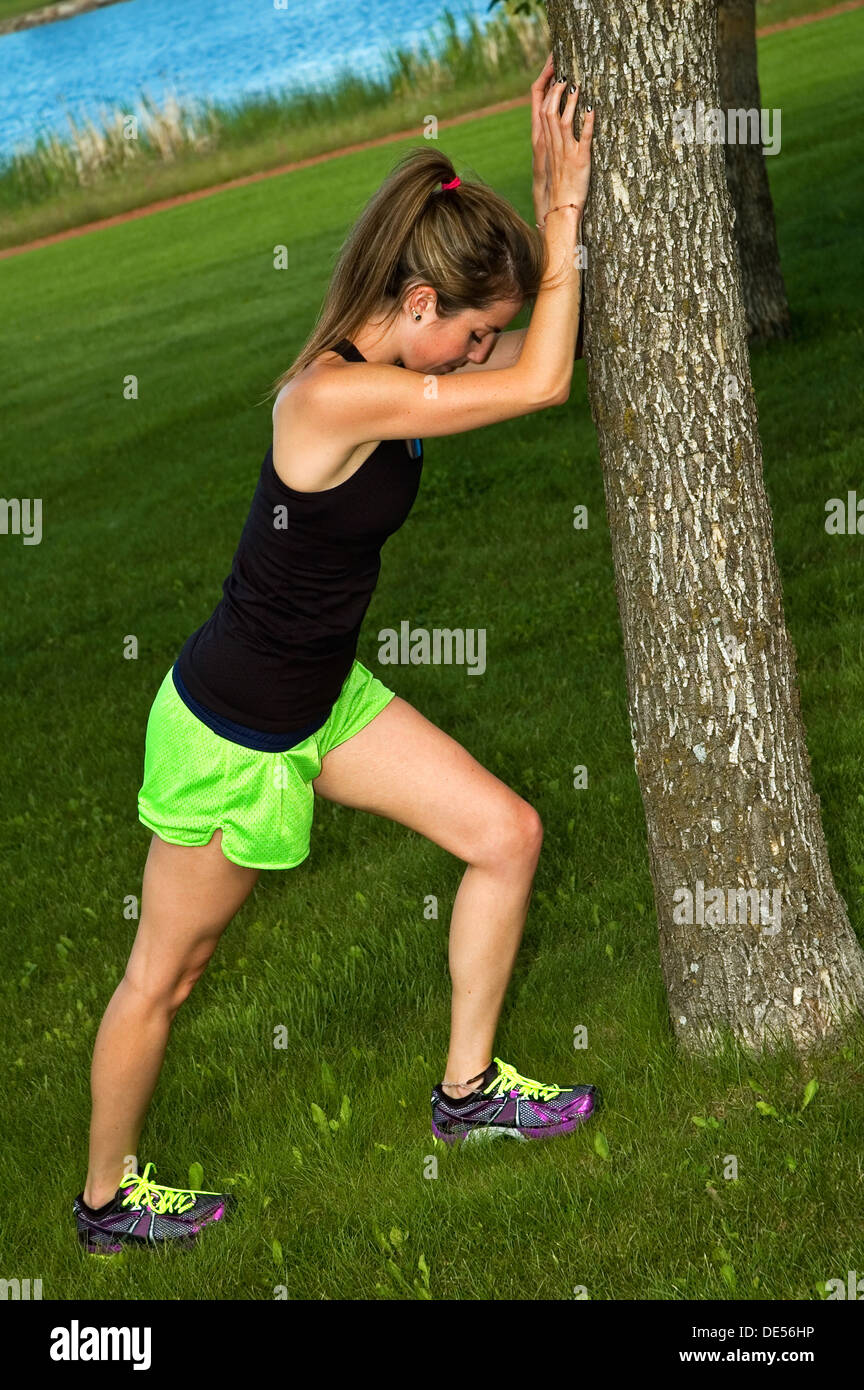 Young woman stretching her calf muscle, against a tree in a city park. Stock Photo