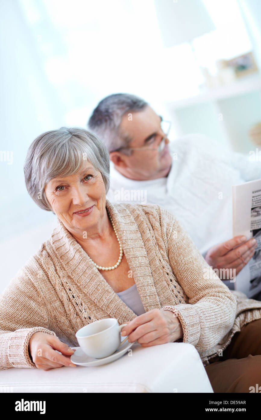 Portrait of mature woman with cup looking at camera on background of her husband reading newspaper Stock Photo