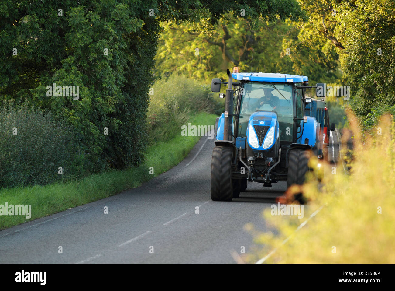 A large tractor travels along a country road taking up a lot of space. Cars can seen seen in the background Stock Photo