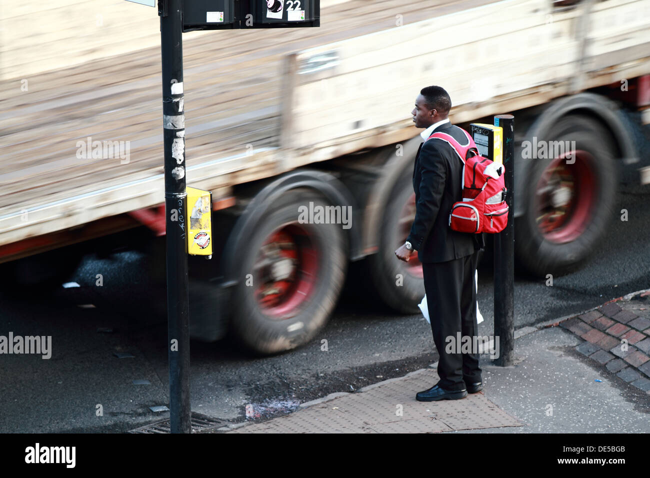 A man stands waiting at the crossing as a large lorry passes closely at speed Stock Photo