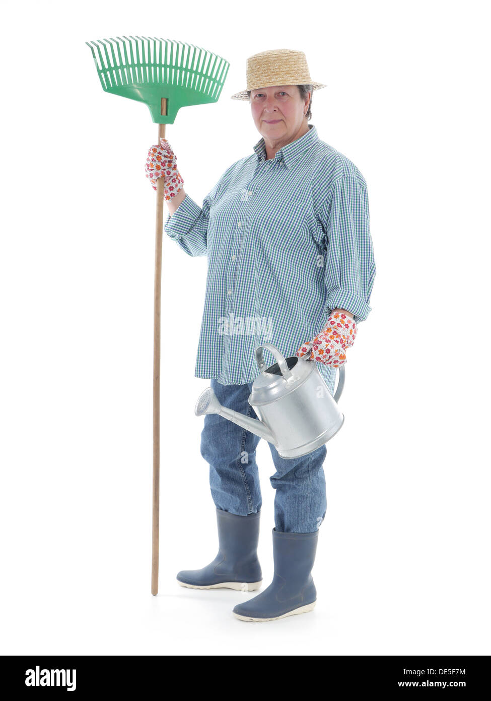 Senior woman gardener wearing straw hat and rubber boots posing with rake and metal watering can over white background Stock Photo