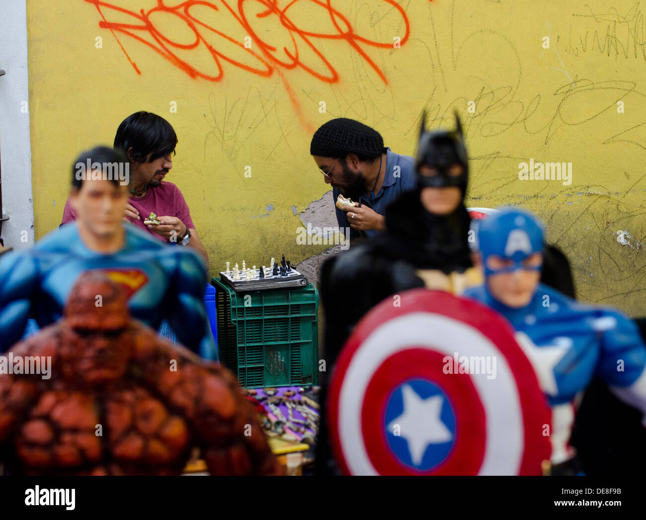 Two men eating snacks and playing chess whilst action superhero figures look on in the foreground. Stock Photo
