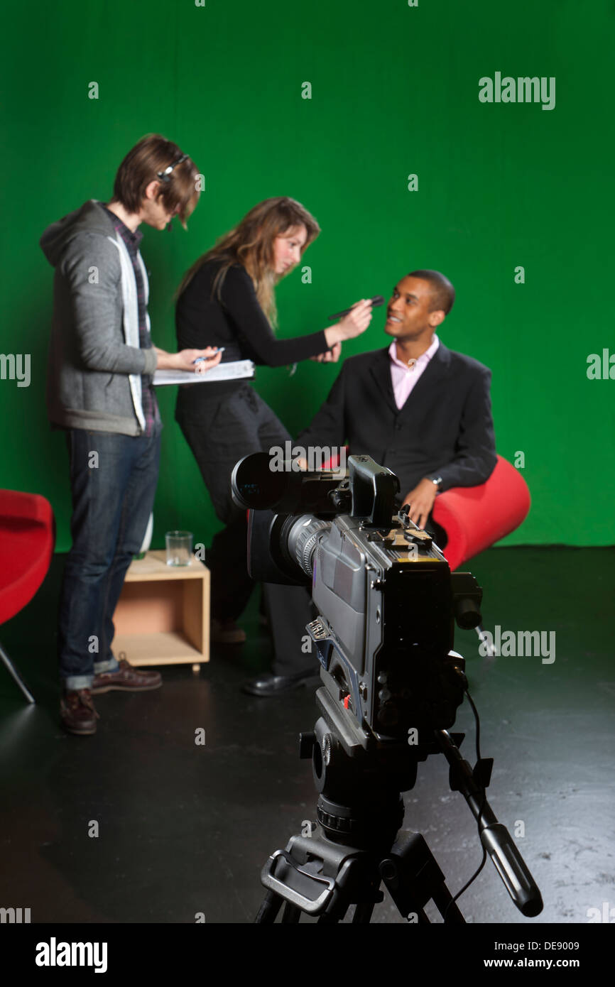 Selective focus on a studio television camera with a floor manager, make-up artist and presenter out of focus in the background. Stock Photo
