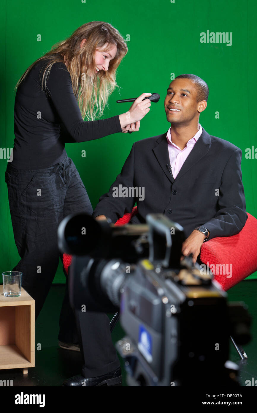 A make-up artist adds final touches to a presenter on a television set with a TV camera out of focus in the foreground. Stock Photo
