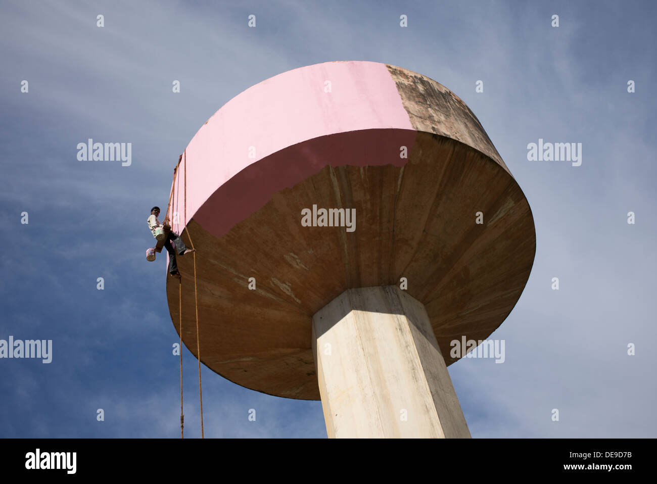 Indian painter sitting on a wooden seat hanging from ropes painting a water tower. Puttaparthi, Andhra Pradesh, India Stock Photo