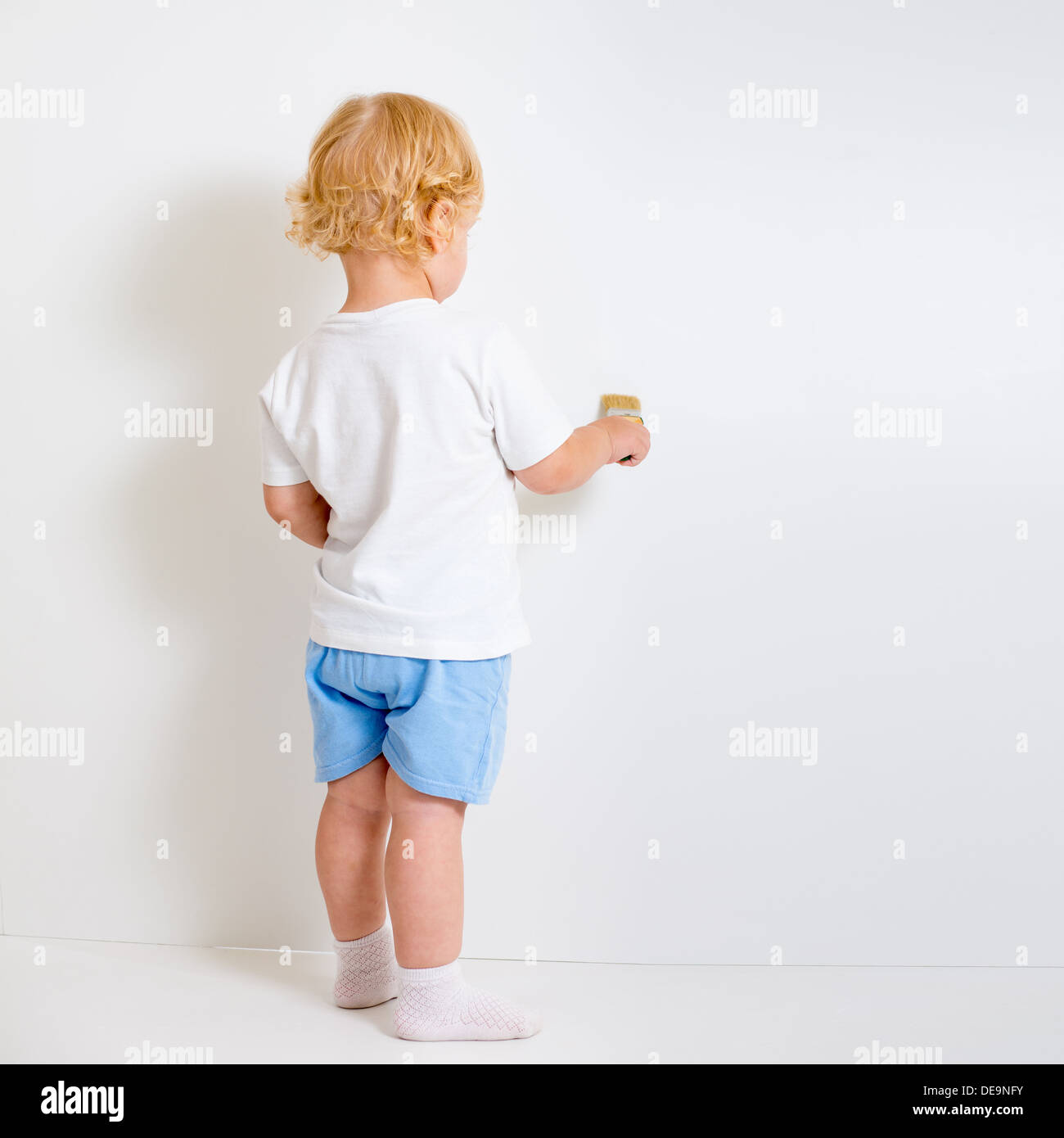 Baby boy with paint brush rear view standing near blank white wall Stock Photo