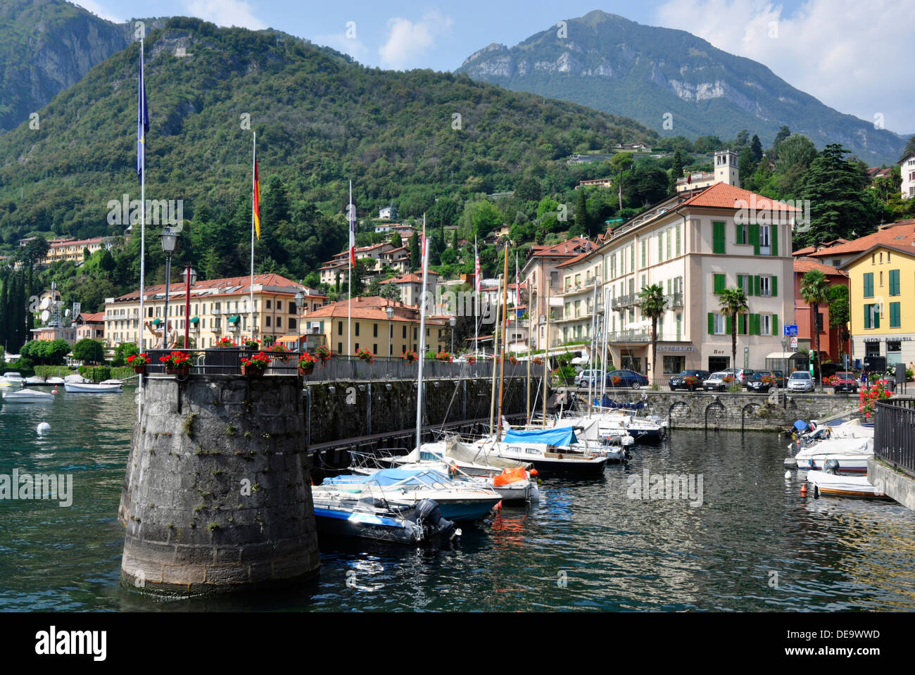 Italy - Lake Como - Menaggio - the harbour - moored yachts - flowers - backdrop town + mountains - sunlight + blue sky Stock Photo