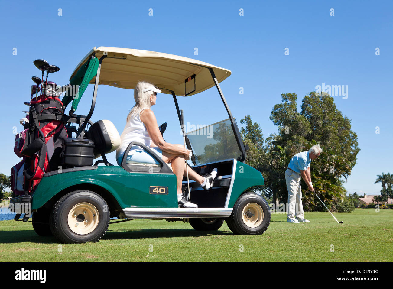 Happy senior man and woman couple together playing golf, the man is hitting a shot the woman is sitting in a golf cart Stock Photo